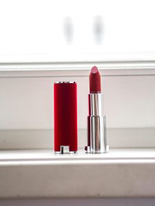 Sculptural gold Valdé Beauty lipstick armor vessel with a red logo inlay representing luxury refillable cosmetics and fine jewelry aesthetic. Close-up of rich, pigmented cream lipstick texture representing contemporary artisanal beauty curation 