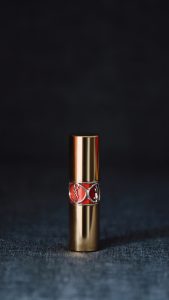 Sculptural gold Valdé Beauty lipstick armor vessel with a red logo inlay representing luxury refillable cosmetics and fine jewelry aesthetic. This captures exactly what is