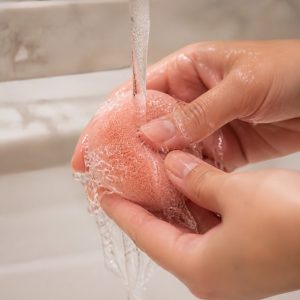 Hands rinsing a pink makeup sponge under running water, part of a clean girl makeup routine care step